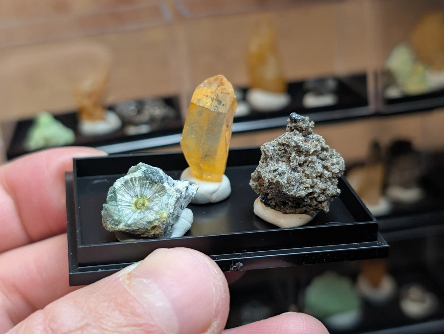 A yellow-orange crystal specimen with a translucent, faceted appearance sits on a white pedestal within a black display tray.