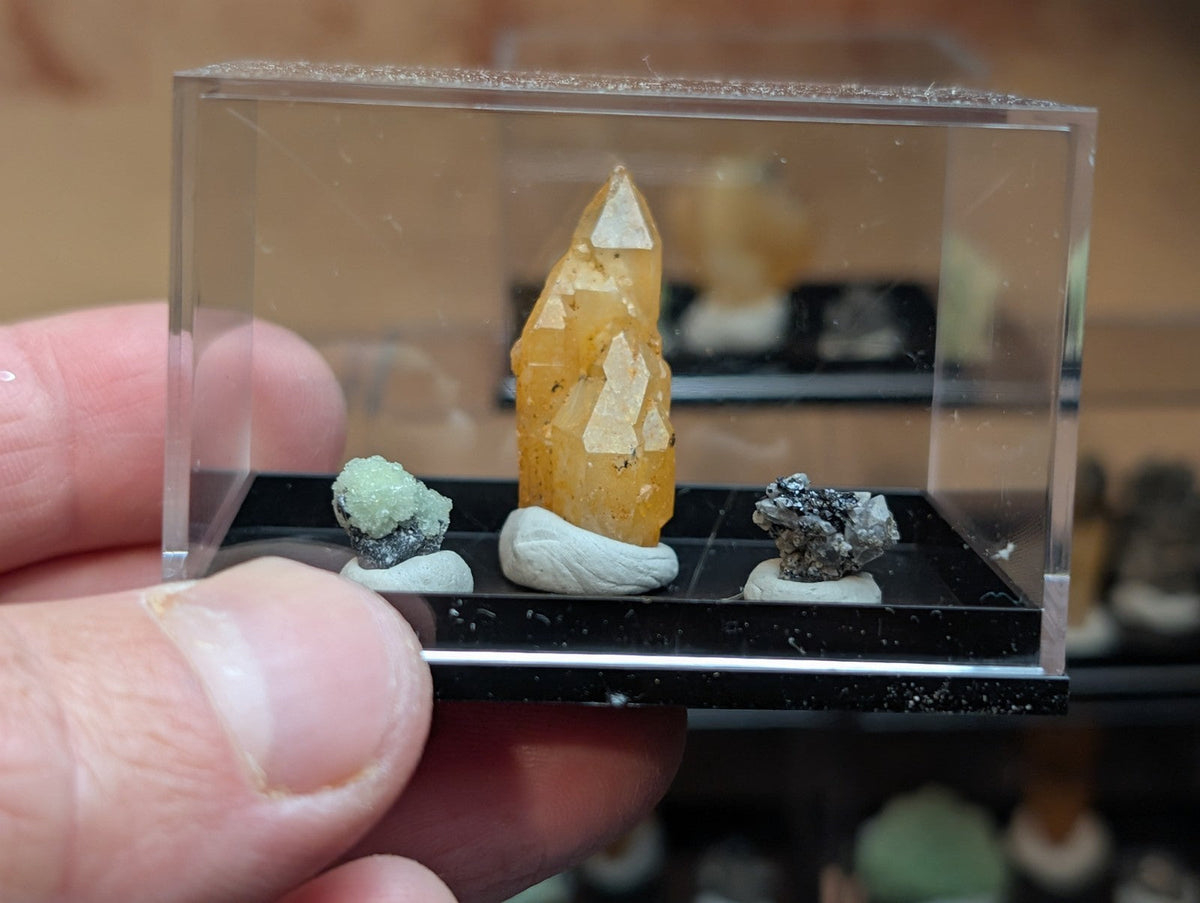 A translucent yellow-orange crystal with white triangular inclusions, displayed upright on a small white pedestal inside a clear acrylic display case.