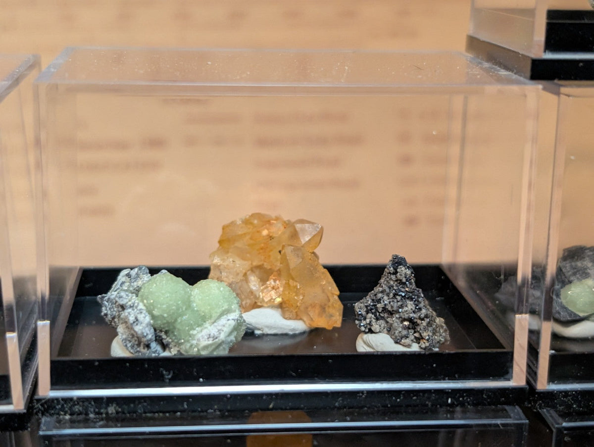 A clear acrylic display case housing three distinct mineral specimens: a cluster of light green crystals with white matrix, an amber-colored translucent crystal formation, and a dark gray-black speckled rock.