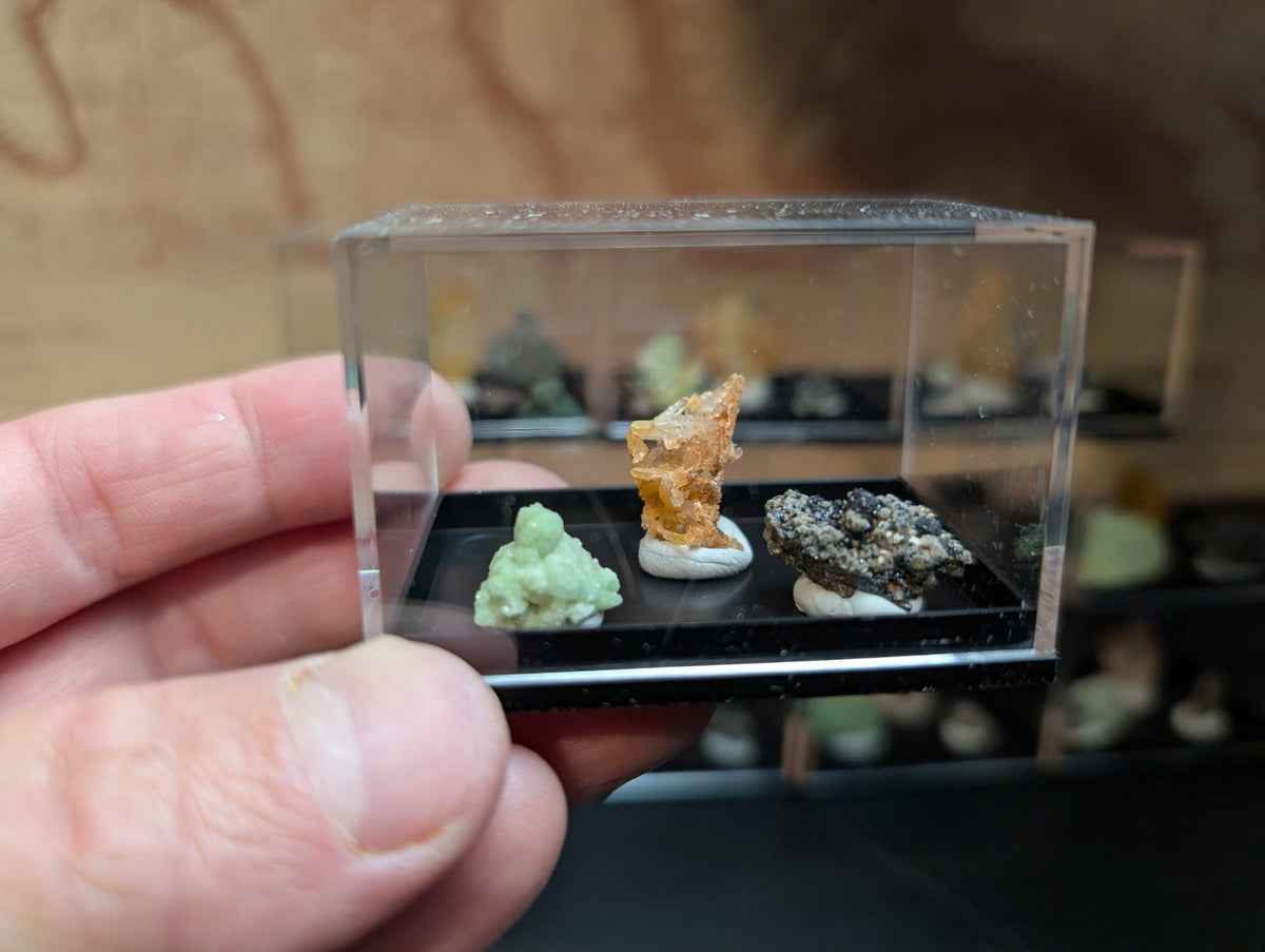 A clear acrylic display case holding three miniature mineral specimens on black bases.