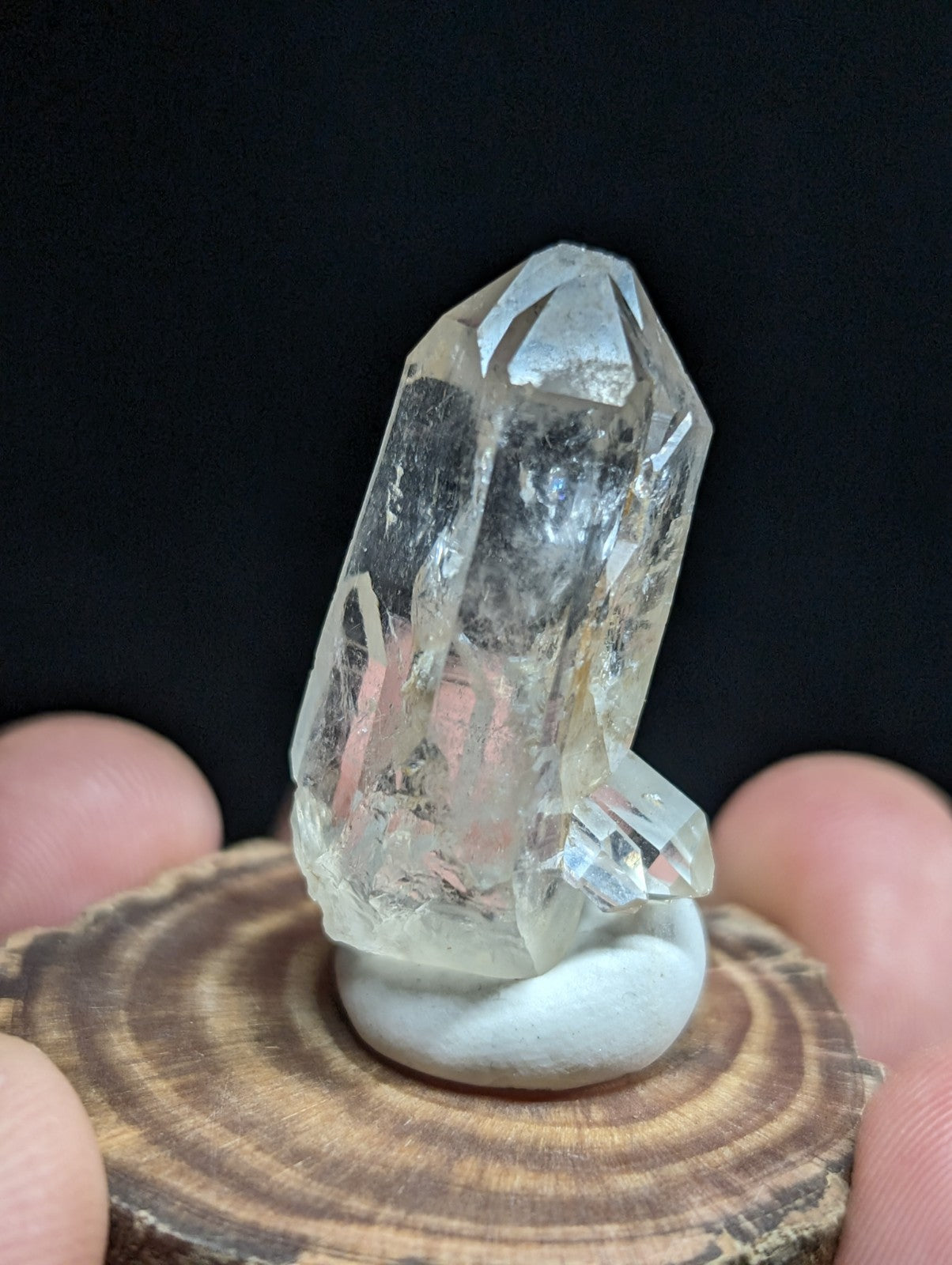A clear, faceted quartz crystal with a pointed tip and natural inclusions, displayed on a white stone base atop a wooden slice.