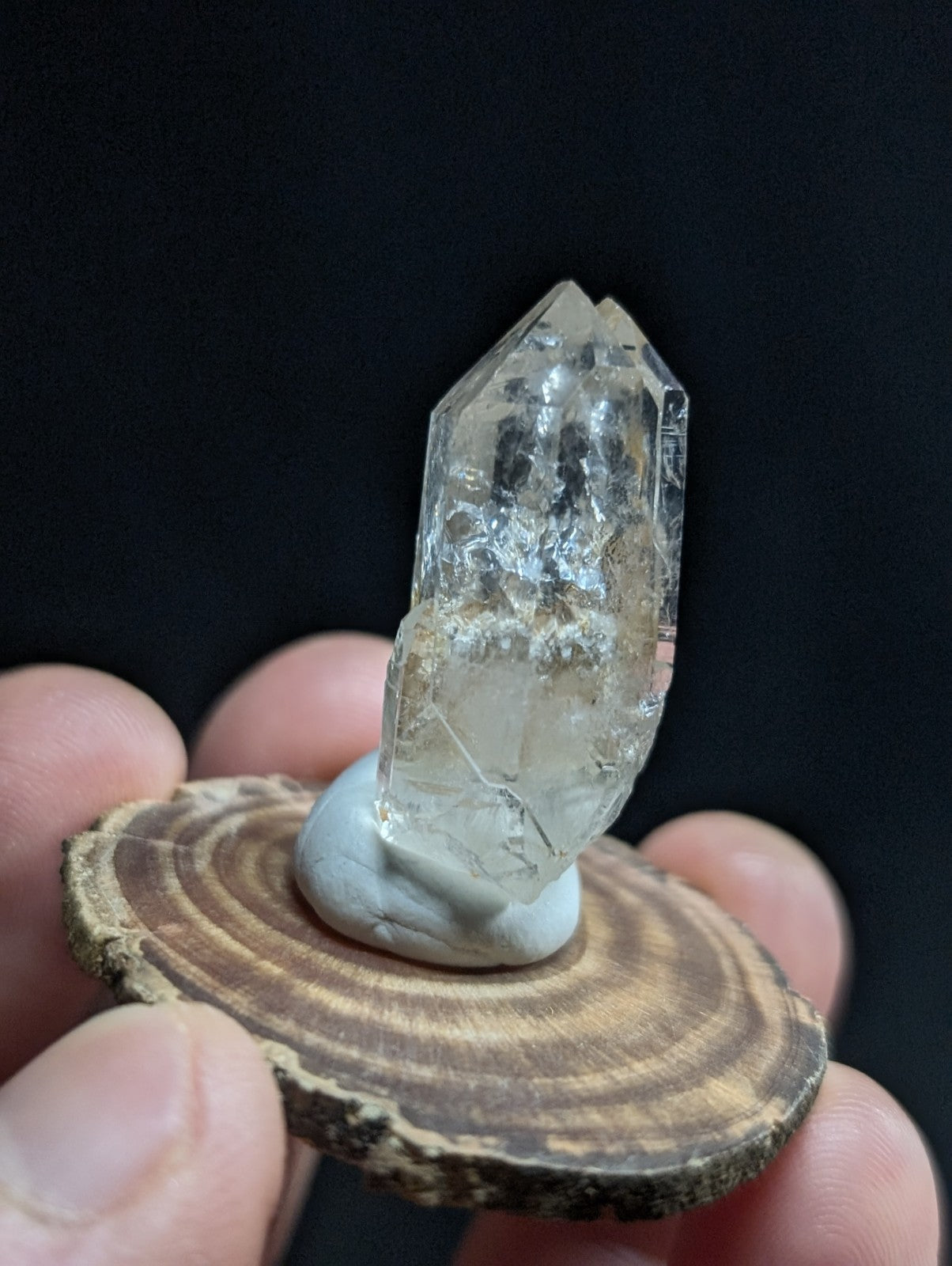 A clear, faceted quartz crystal with natural inclusions sits atop a smooth white base on a rustic wooden slice.
