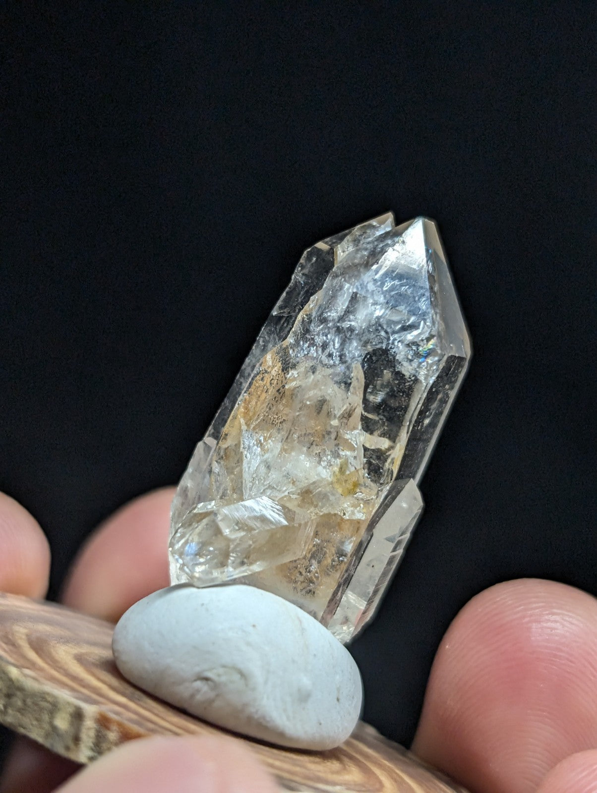 A clear, faceted quartz crystal with subtle golden inclusions rests on a smooth white stone atop a wooden surface.