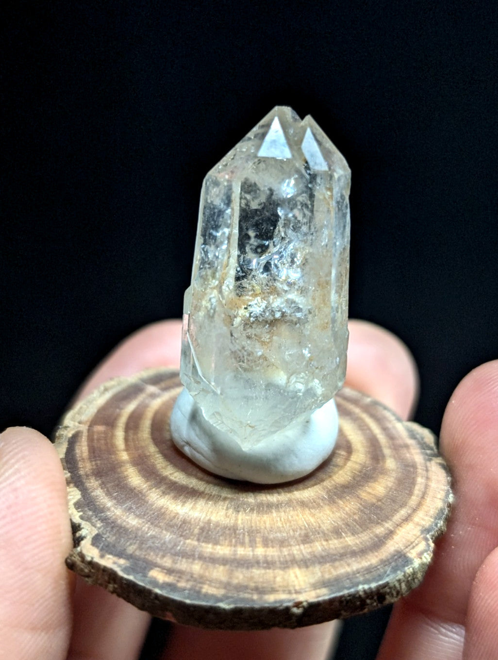 A clear quartz crystal with a pointed, faceted top and natural inclusions sits on a white base atop a circular wooden slice.