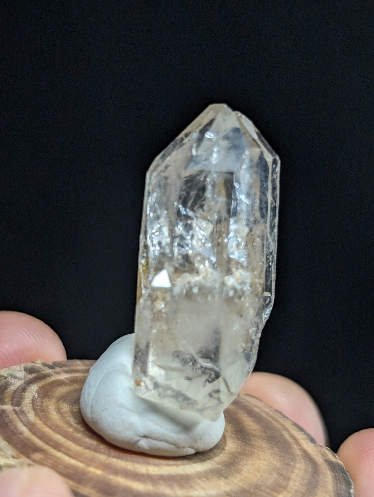A clear, faceted quartz crystal with natural inclusions and a pointed tip, displayed on a smooth white base atop a wooden slice.
