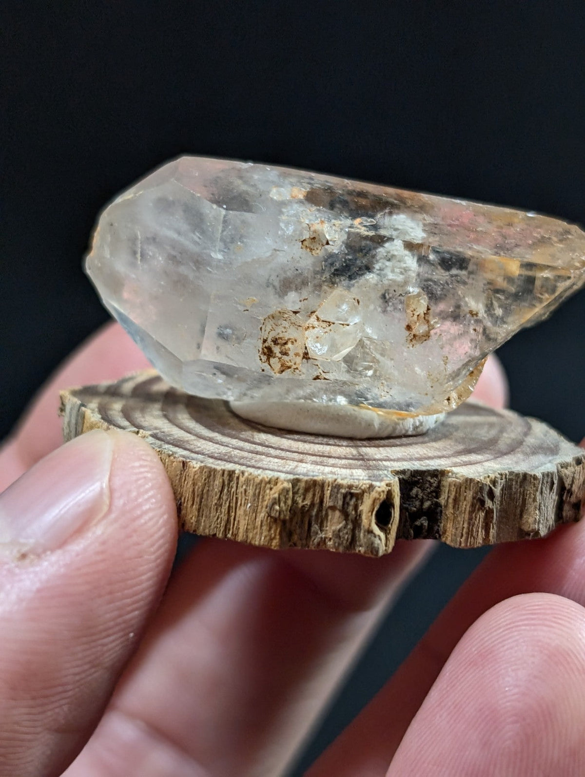 A translucent, faceted quartz crystal with natural inclusions and a slightly amber tint rests on a rustic wooden slice.