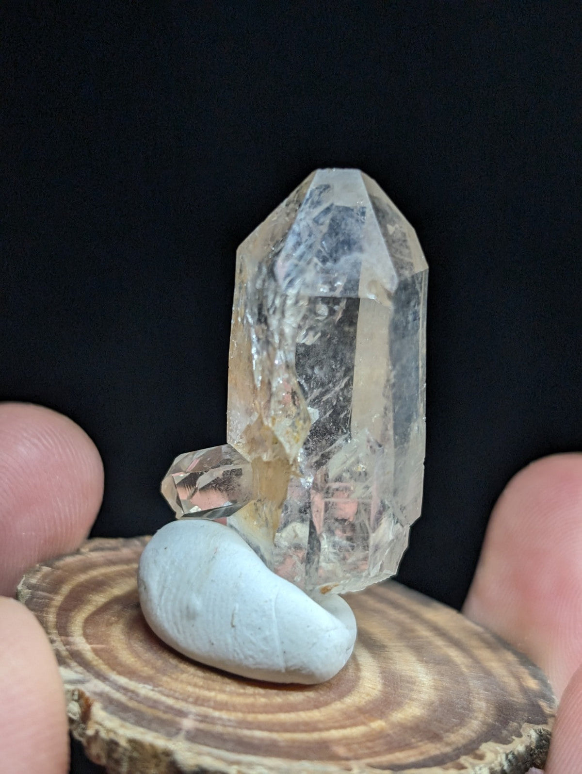 A clear, faceted quartz crystal with a yellowish tint stands upright on a smooth, light blue stone atop a circular wooden slice.