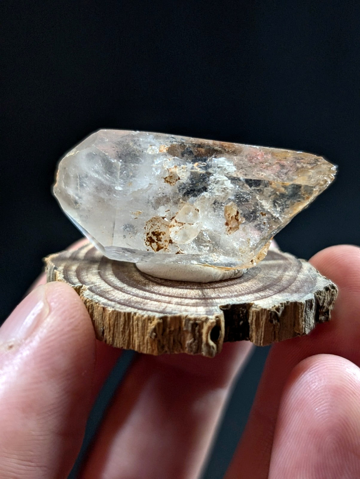 A translucent, clear quartz crystal with natural inclusions and earthy brown mineral deposits rests on a rustic wooden slice.