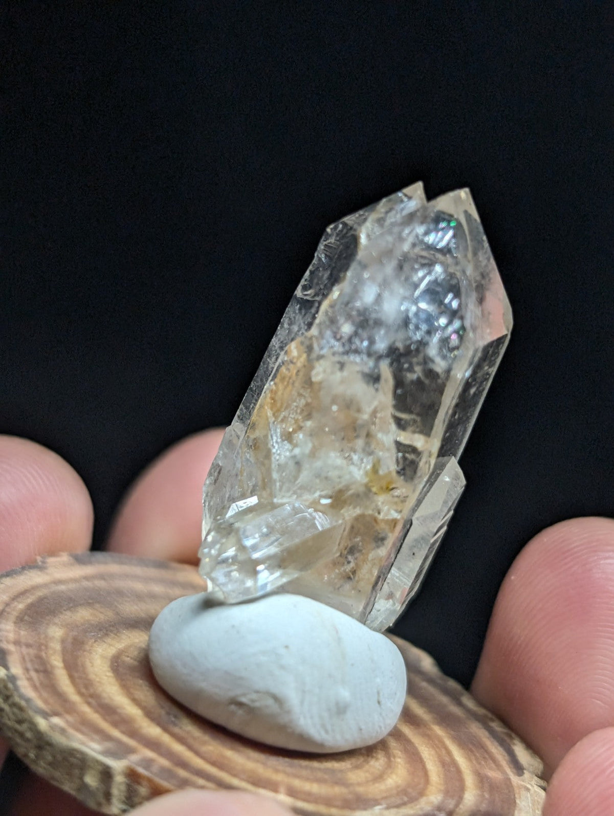 A translucent, faceted quartz crystal with subtle golden inclusions rests on a smooth white stone atop a circular wooden slice.