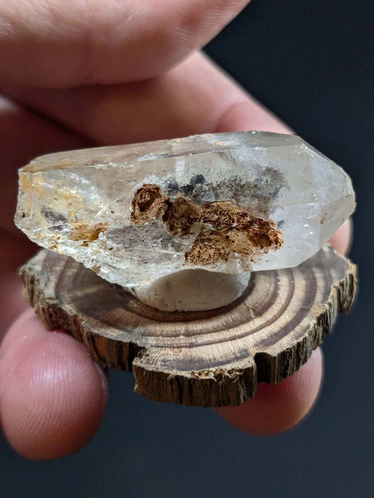 A translucent quartz crystal with brown and black inclusions, held above a circular wooden slice.