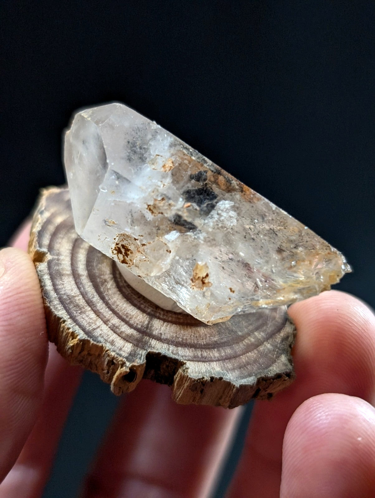 A translucent, faceted quartz crystal with natural inclusions and earthy brown markings rests on a rustic wooden slice.