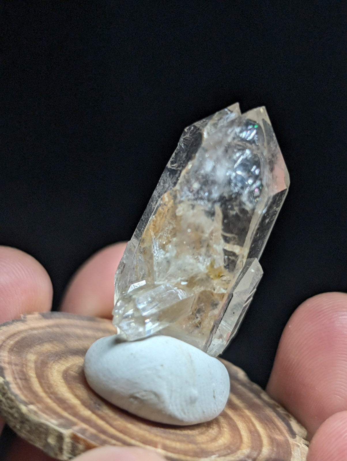 A translucent, faceted quartz crystal with subtle golden inclusions rests on a smooth white stone atop a circular wooden slice.
