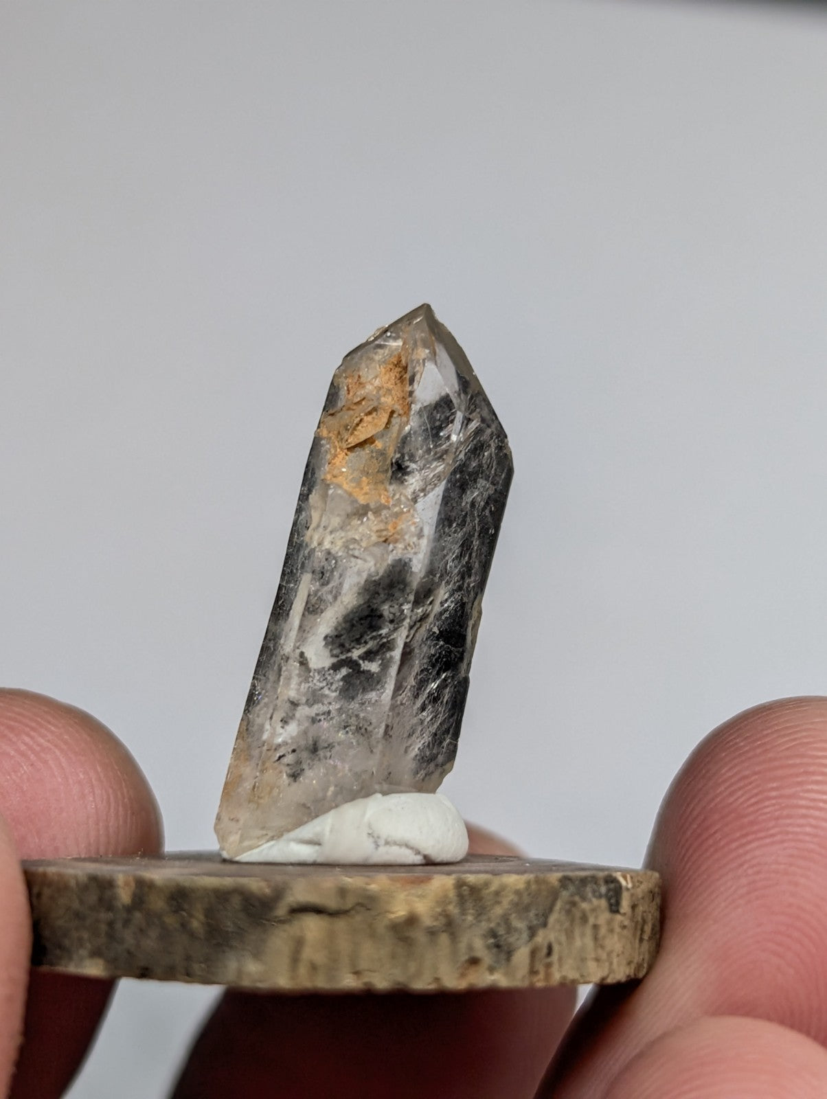 A translucent quartz crystal with dark inclusions and a golden mineral deposit, held upright on a rustic wooden base.