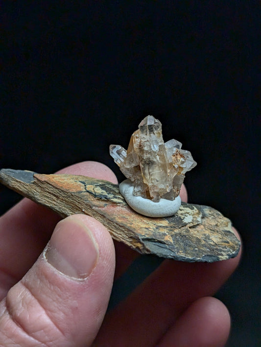 A translucent, crystalline quartz cluster with a natural amber hue rests atop a smooth white stone, which is set upon an irregularly shaped piece of earthy-toned rock held gently in someone’s hand.