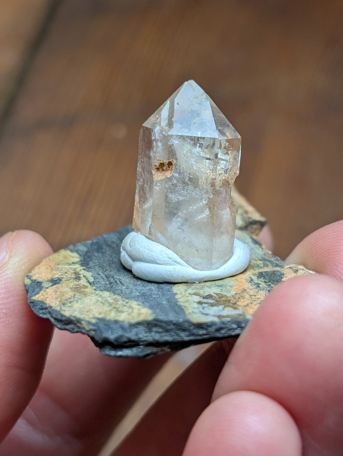 A clear quartz crystal with a pointed tip, mounted on a small piece of dark rock and held in someone’s hand.