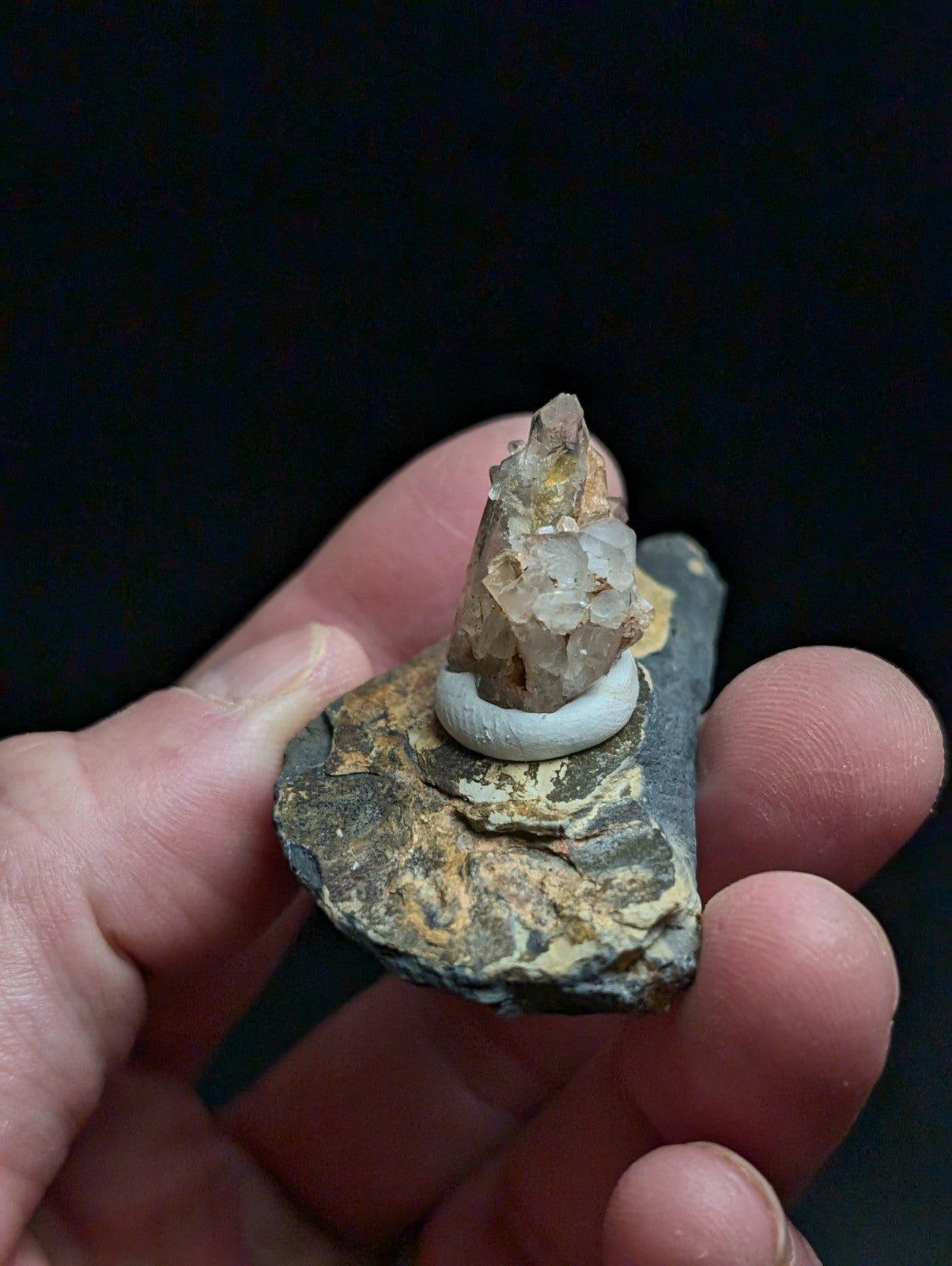 A translucent, crystalline quartz point with golden inclusions sits atop a white ceramic ring, mounted on a dark gray rock base.
