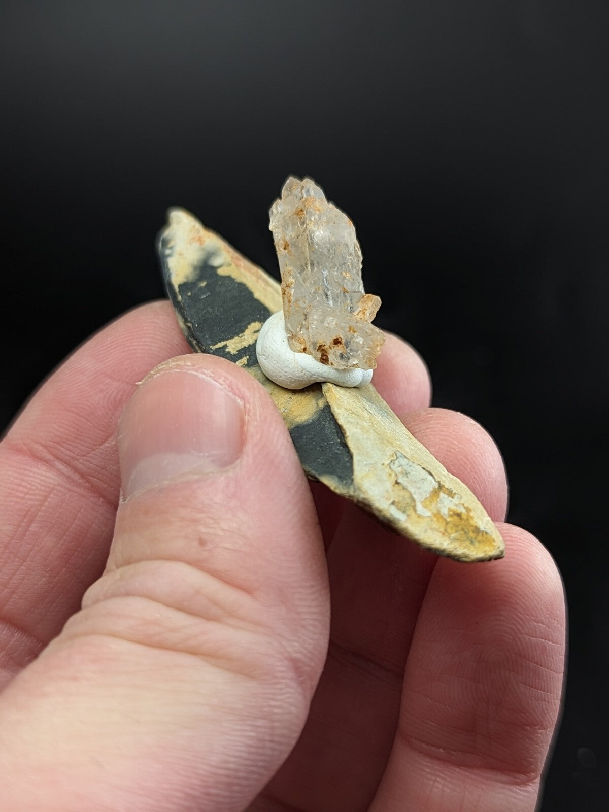 A hand holds a weathered, dark green and yellow ceramic shard with a clear quartz crystal mounted on it using white adhesive.
