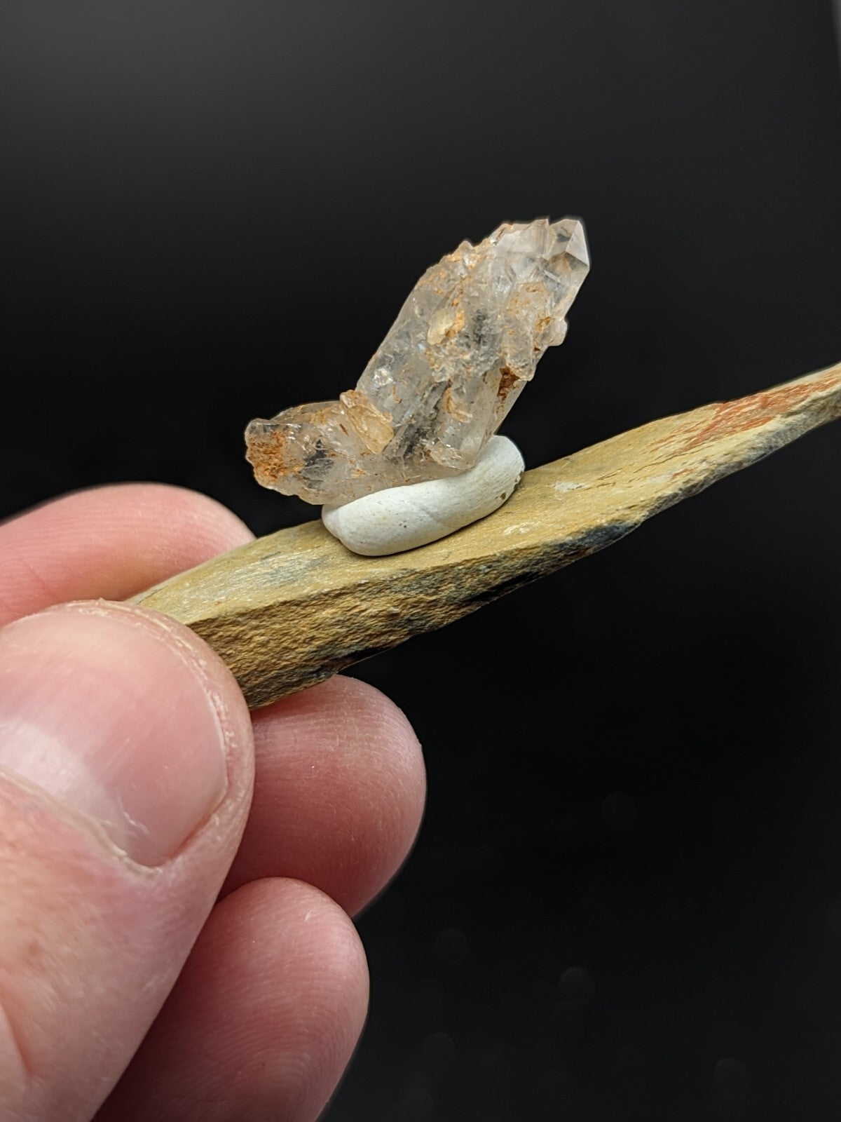 A translucent, faceted quartz crystal with golden inclusions rests atop a smooth white stone on a slender, textured wooden stick.