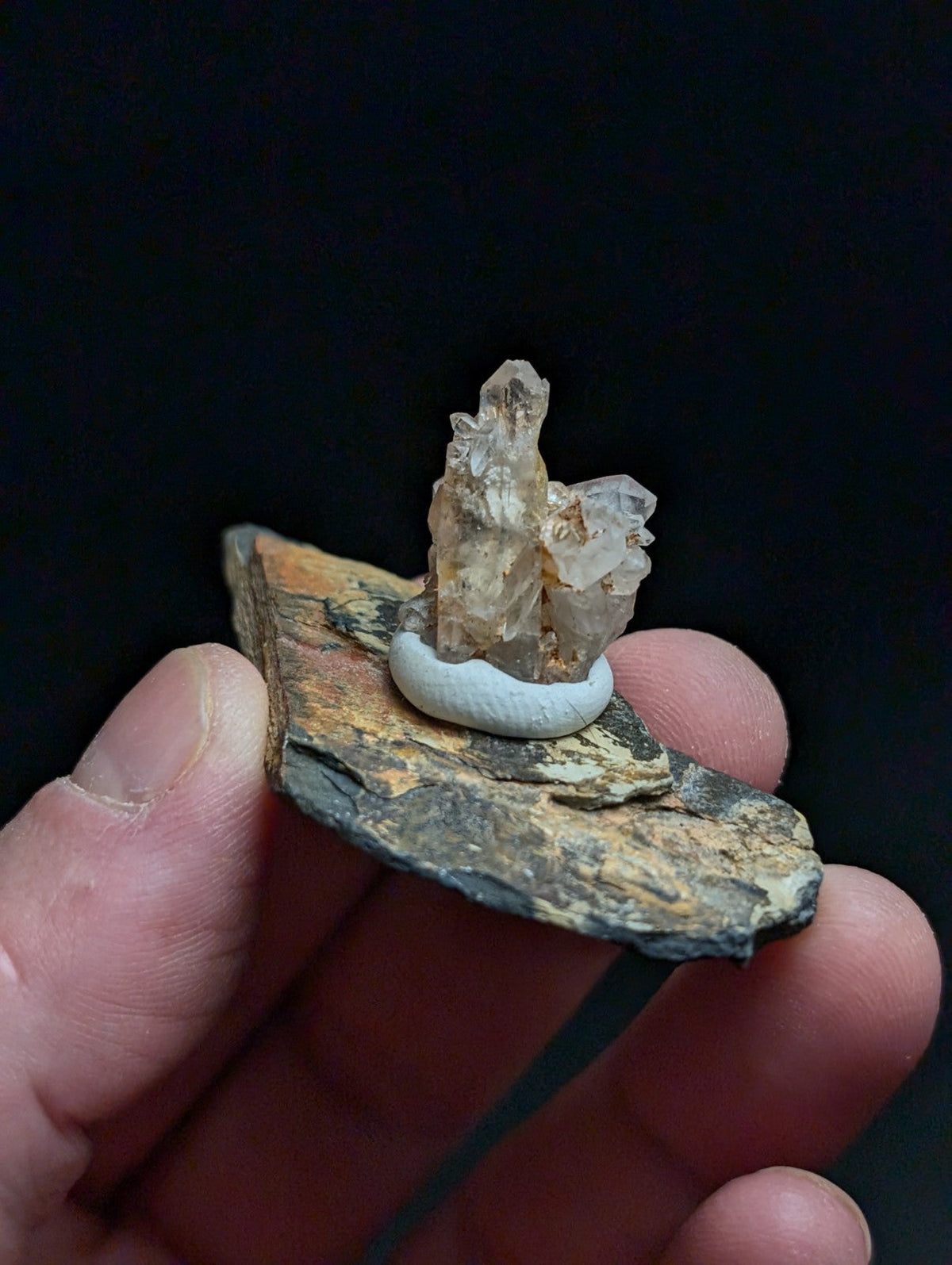 A translucent, multi-faceted quartz crystal cluster rests atop a smooth white circular base, which is set upon a rugged, earth-toned rock slab.