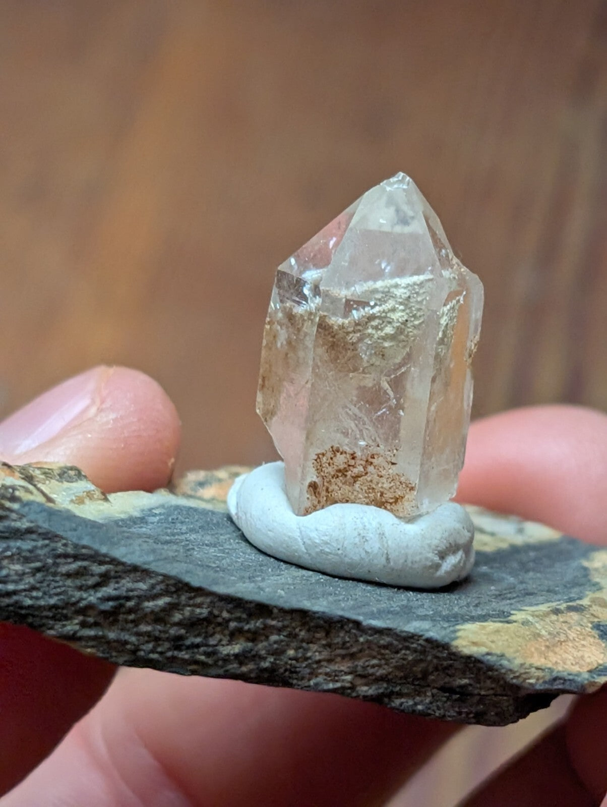 A clear, faceted quartz crystal with golden inclusions sits atop a smooth white base on a dark stone slab.