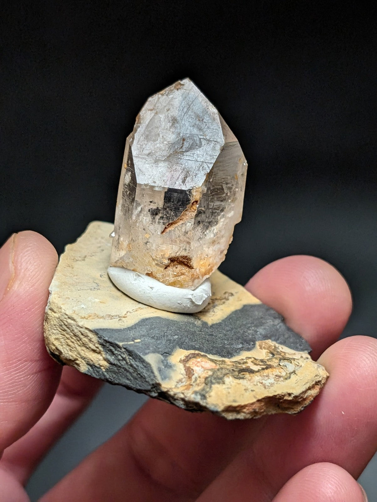 A translucent, faceted quartz crystal with subtle brown inclusions sits atop a white circular base mounted on a layered rock slab.