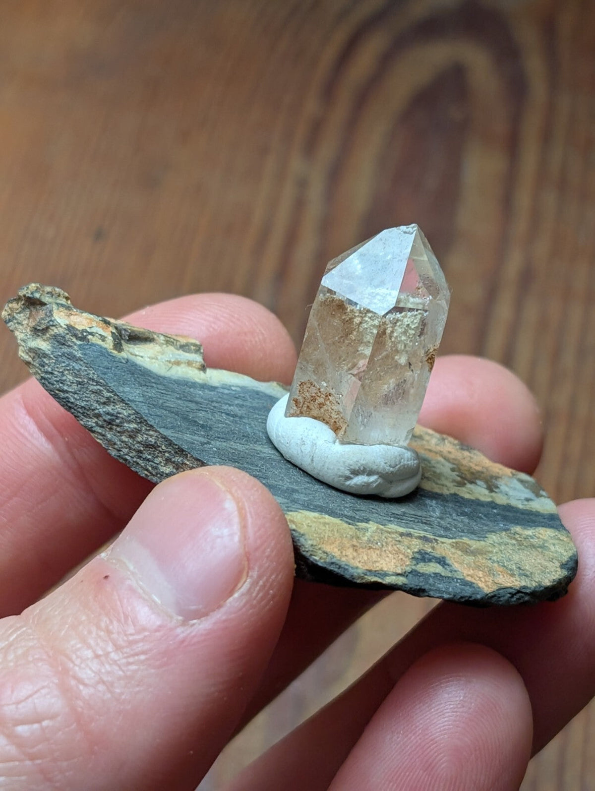 A clear, faceted quartz crystal with a slightly cloudy base sits atop a small white stone, which is placed on a flat, dark gray and brown striped rock slab.
