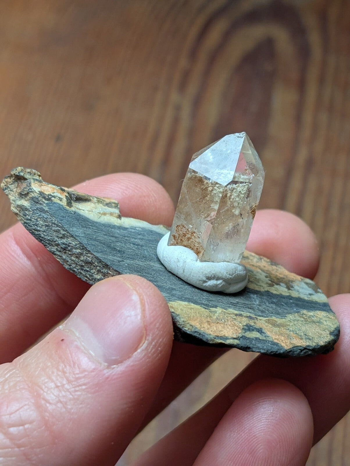 A clear, faceted quartz crystal with a slightly cloudy base sits atop a small white stone, which is placed on a flat, dark gray and brown striped rock slab.