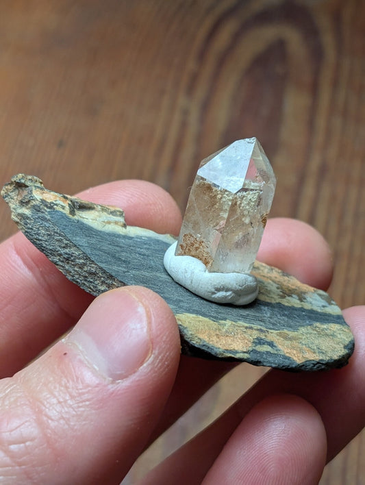 A clear, faceted quartz crystal with a slightly cloudy base sits atop a small white stone, which is placed on a flat, dark gray and brown striped rock slab.