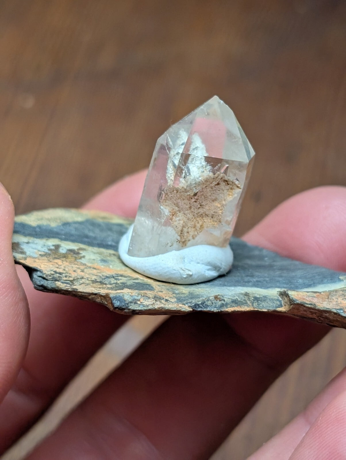 A clear quartz crystal with a natural, earthy inclusion sits elegantly on a small white pedestal atop a flat, textured stone slab.