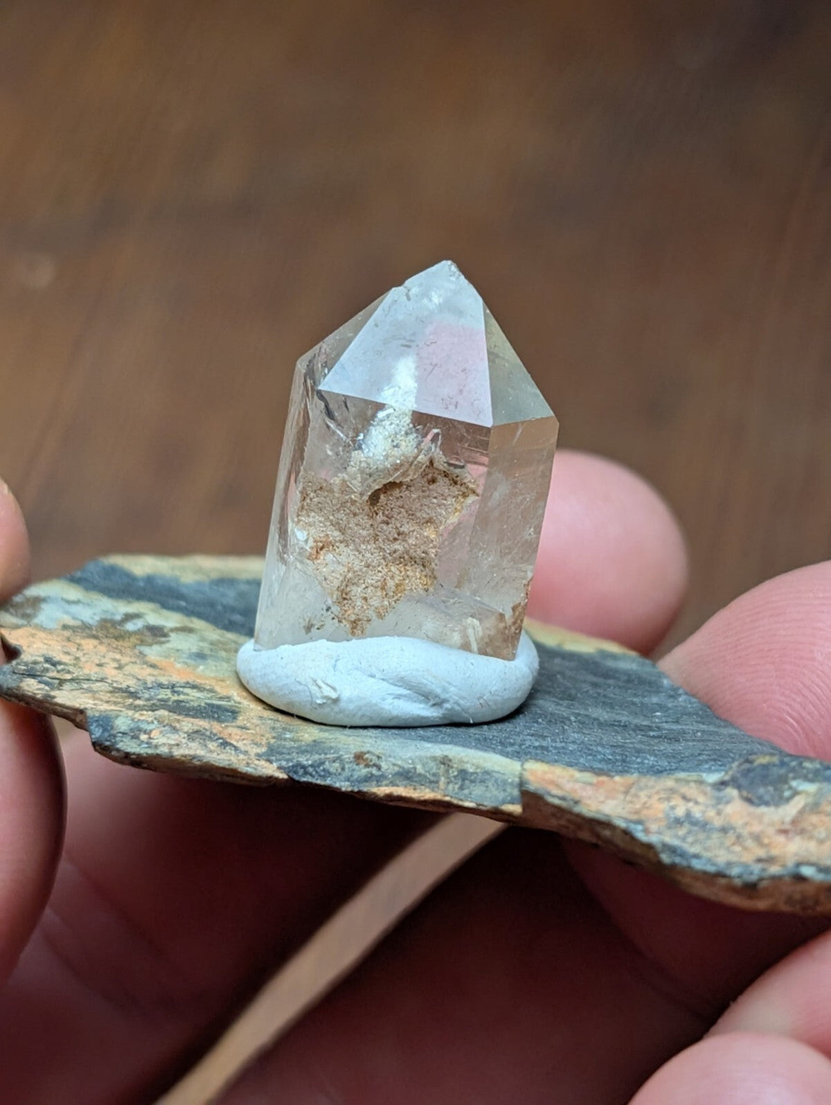 A clear quartz crystal with a pointed tip and internal earthy inclusions, mounted on a light-colored base atop a dark, textured stone slab.