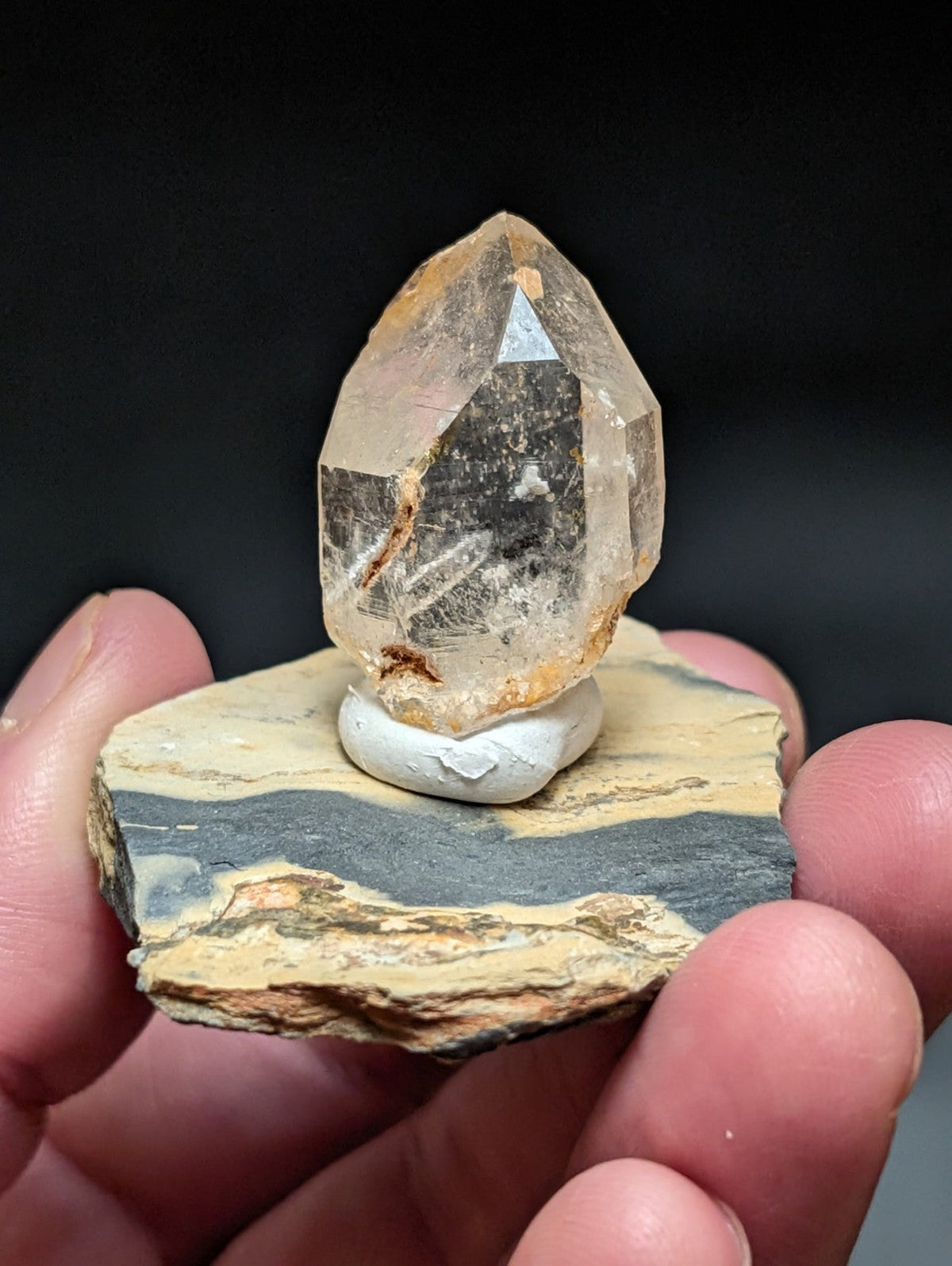 A translucent, faceted citrine crystal with golden-brown inclusions sits atop a white ceramic base mounted on a layered rock slab.