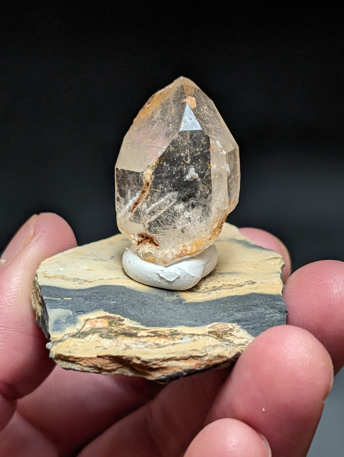 A translucent, faceted citrine crystal with golden-brown inclusions sits atop a white ceramic base mounted on a layered rock slab.