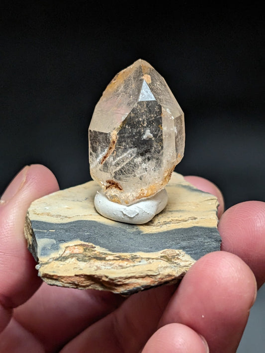 A translucent, faceted citrine crystal with golden-brown inclusions sits atop a white ceramic base mounted on a layered rock slab.