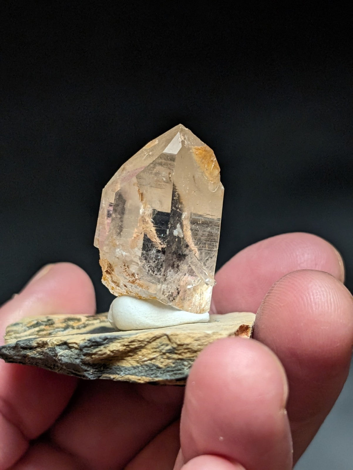 A translucent, amber-hued quartz crystal with natural inclusions and a pointed tip, displayed on a small white pedestal atop a textured rock base.