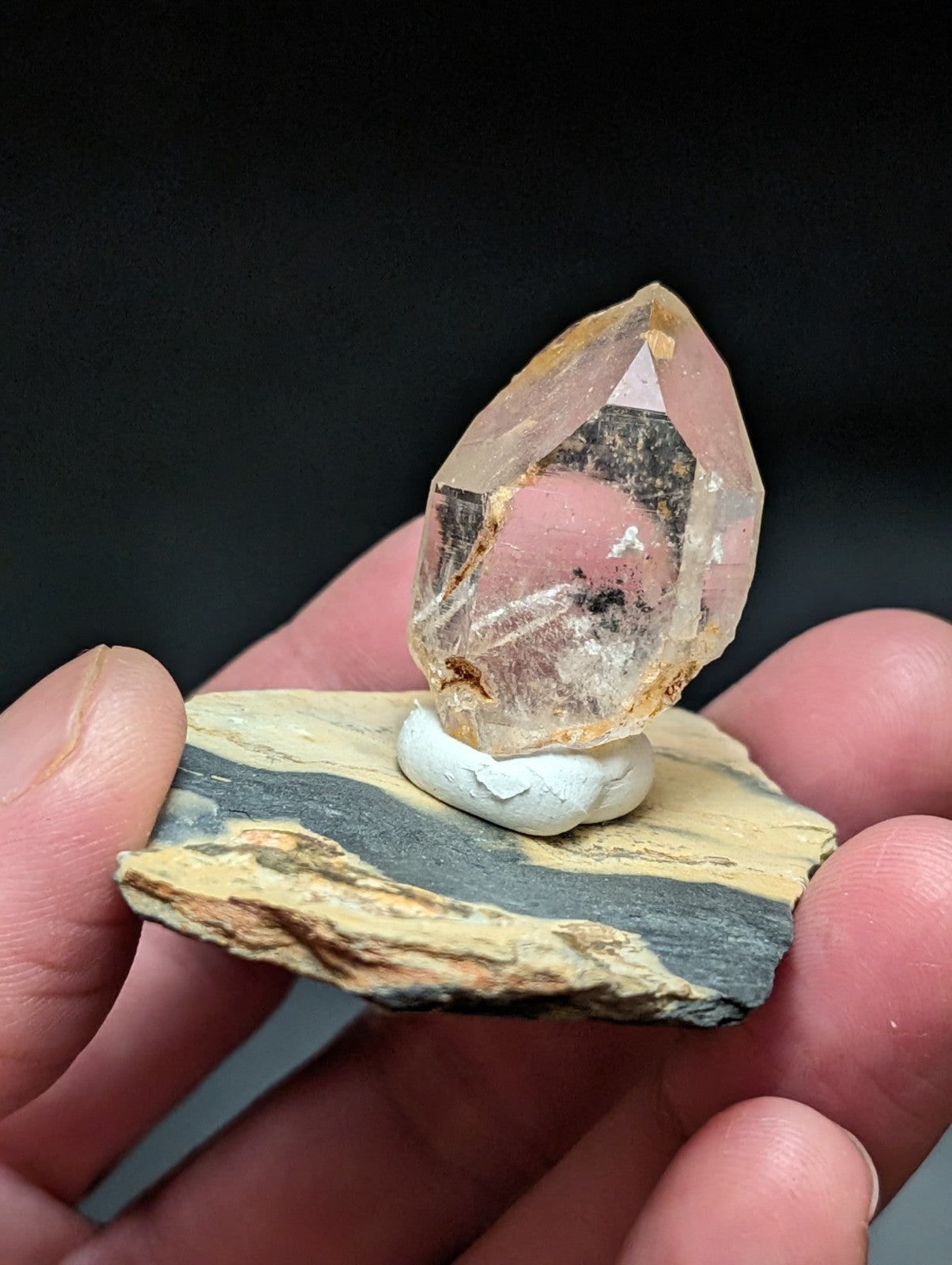 A translucent, pale yellowish crystal with a pointed tip and internal inclusions sits atop a small white pedestal on a layered rock slab.