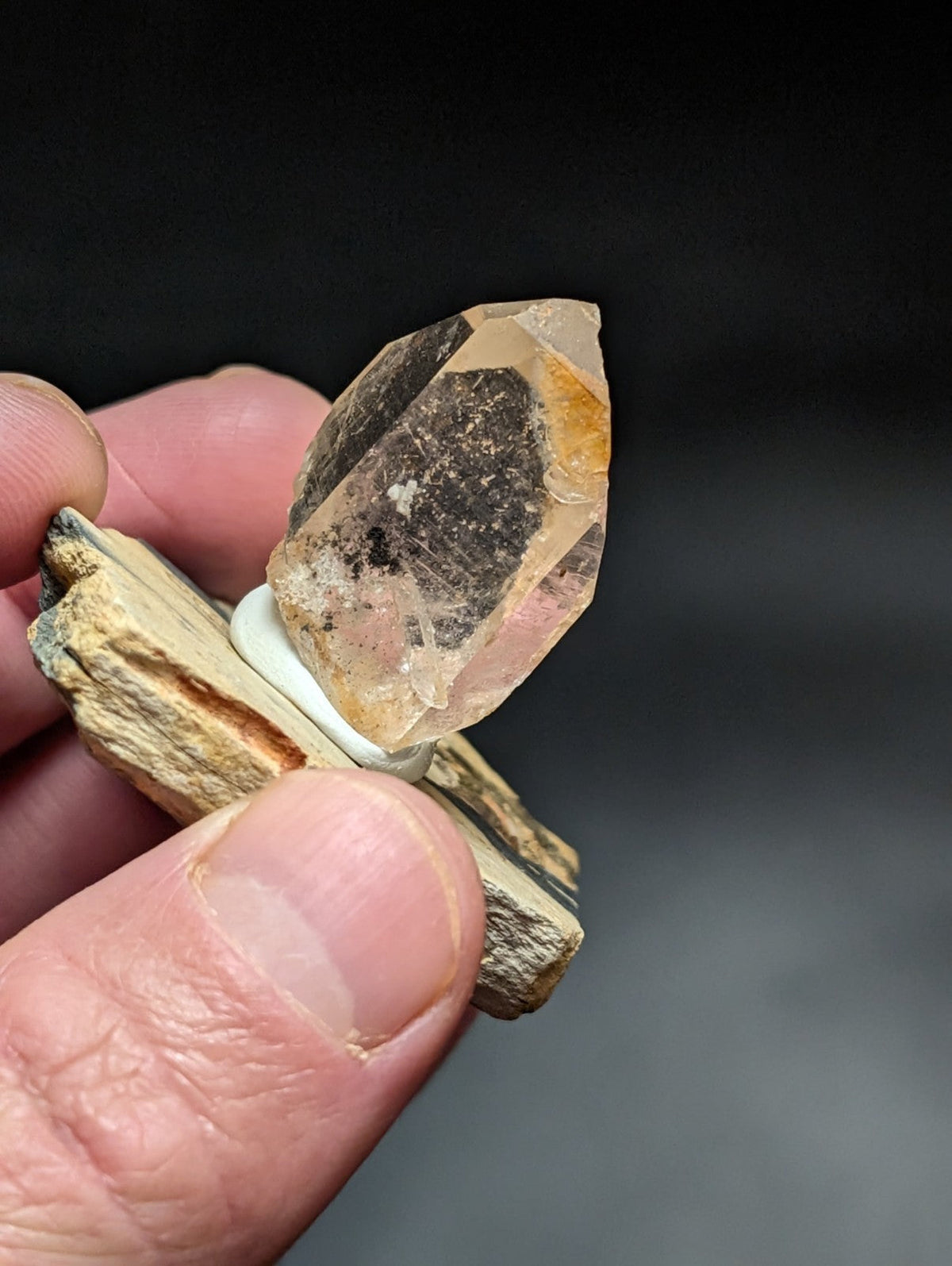 A translucent, amber-hued quartz crystal with a dark inclusions and sharp facets is held between fingers.