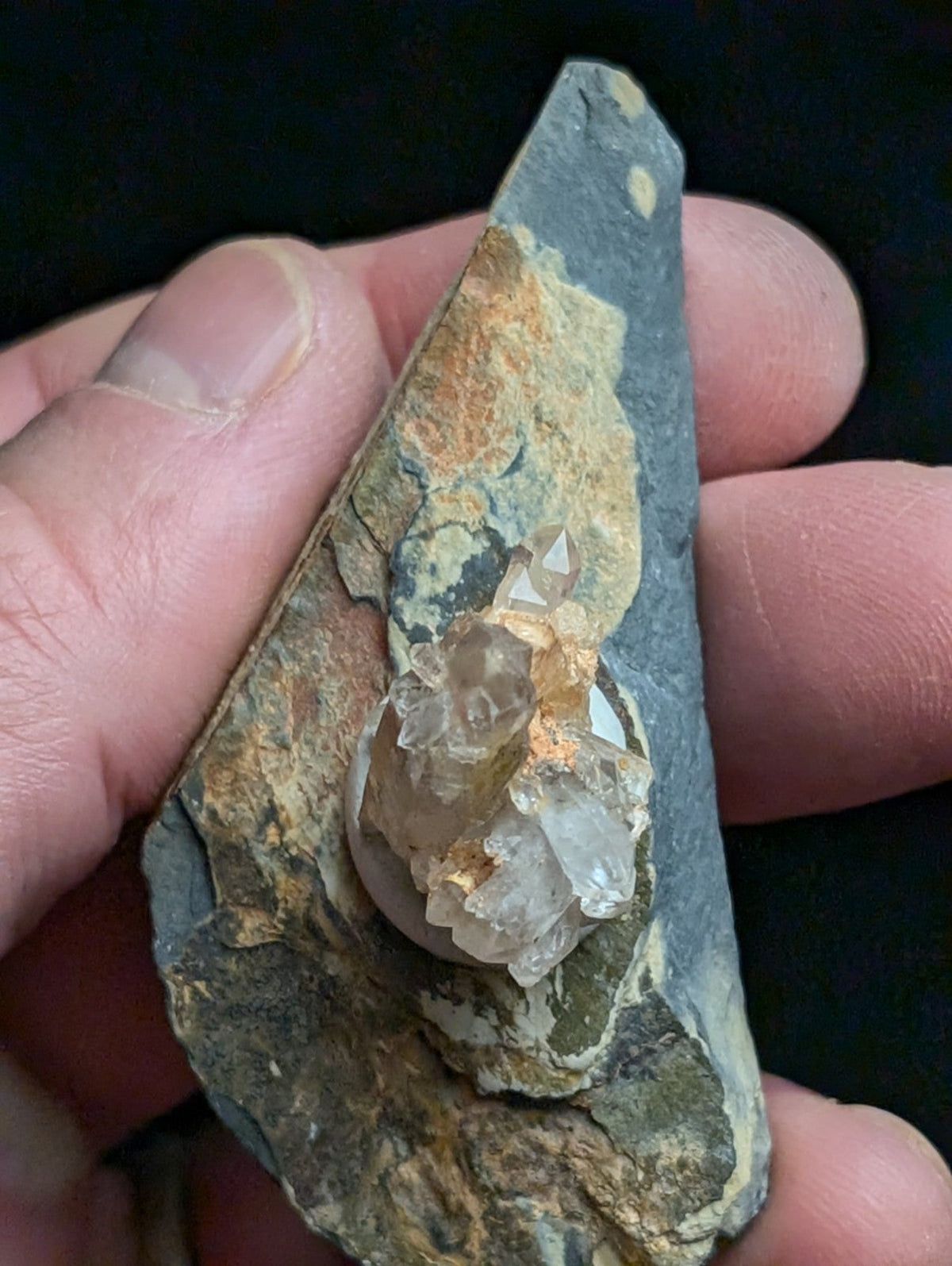 A cluster of translucent, crystalline quartz formations nestled within a dark gray rock matrix with patches of orange and green mineralization.