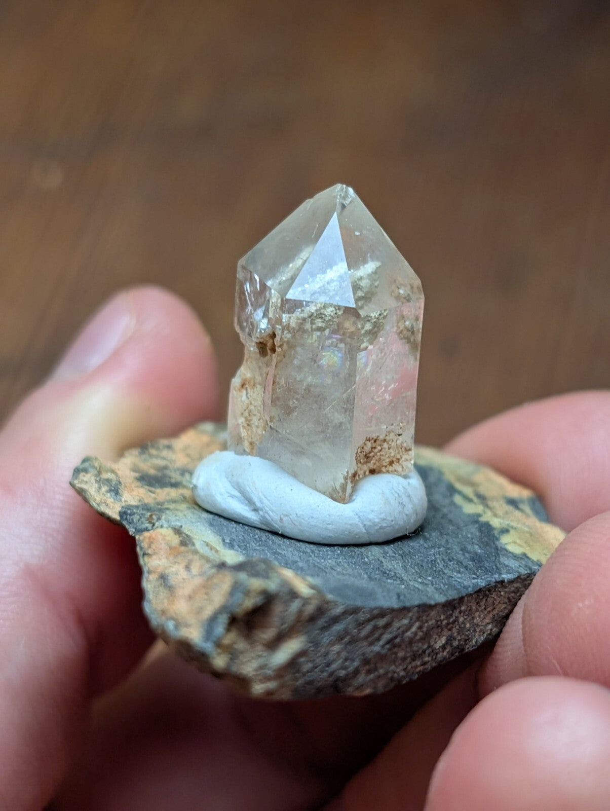 A clear, faceted quartz crystal with natural inclusions sits atop a small white clay base mounted on a dark, textured rock.