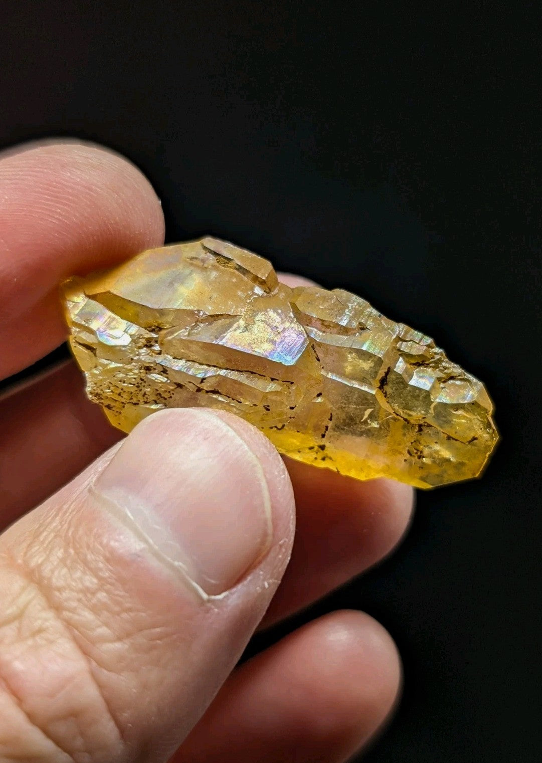 A translucent golden-yellow crystal with iridescent facets and natural mineral inclusions, held gently between fingers.