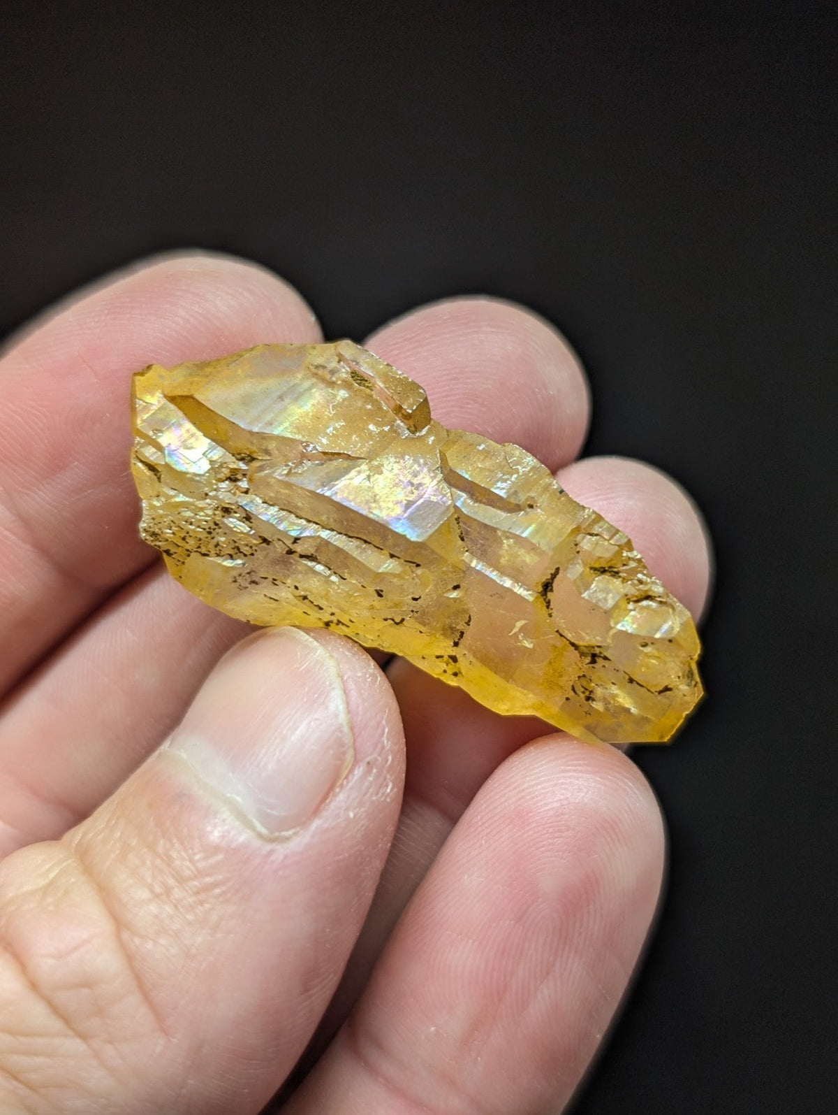 A translucent golden-yellow crystal with iridescent facets and dark inclusions, held gently between fingers.