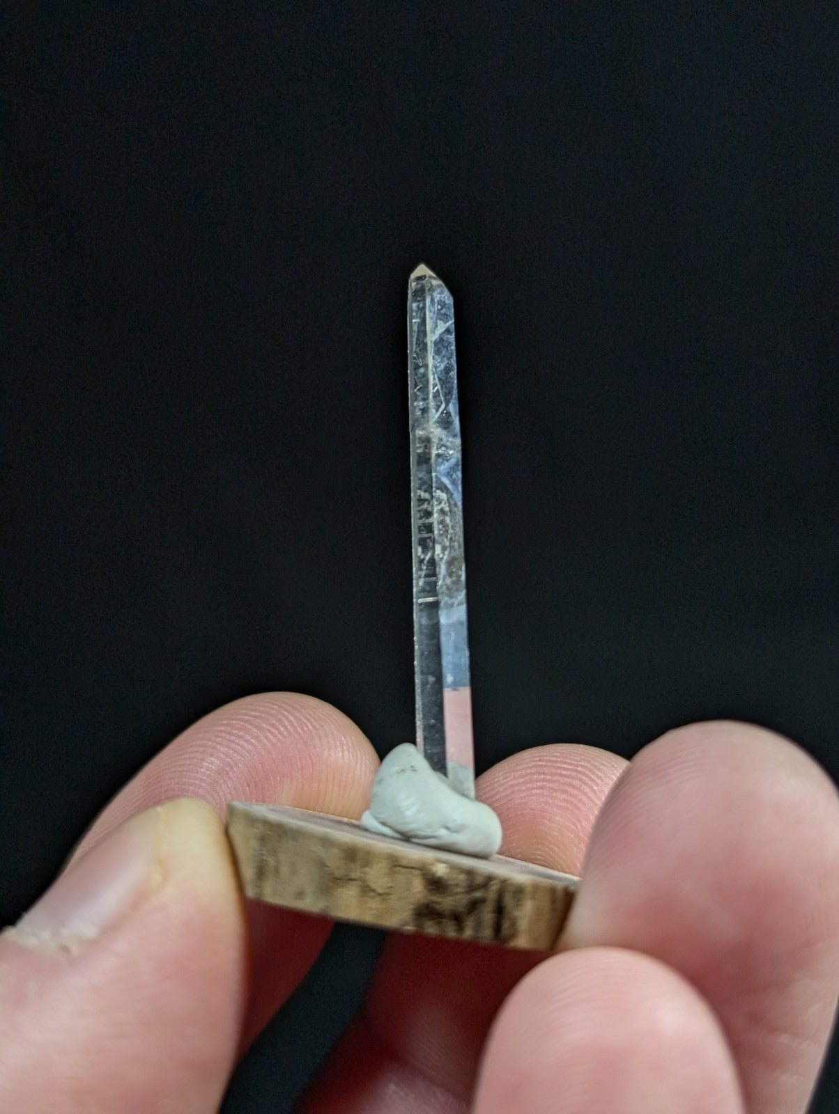 A clear, faceted quartz crystal with subtle internal veining stands upright on a small wooden base held between fingers.