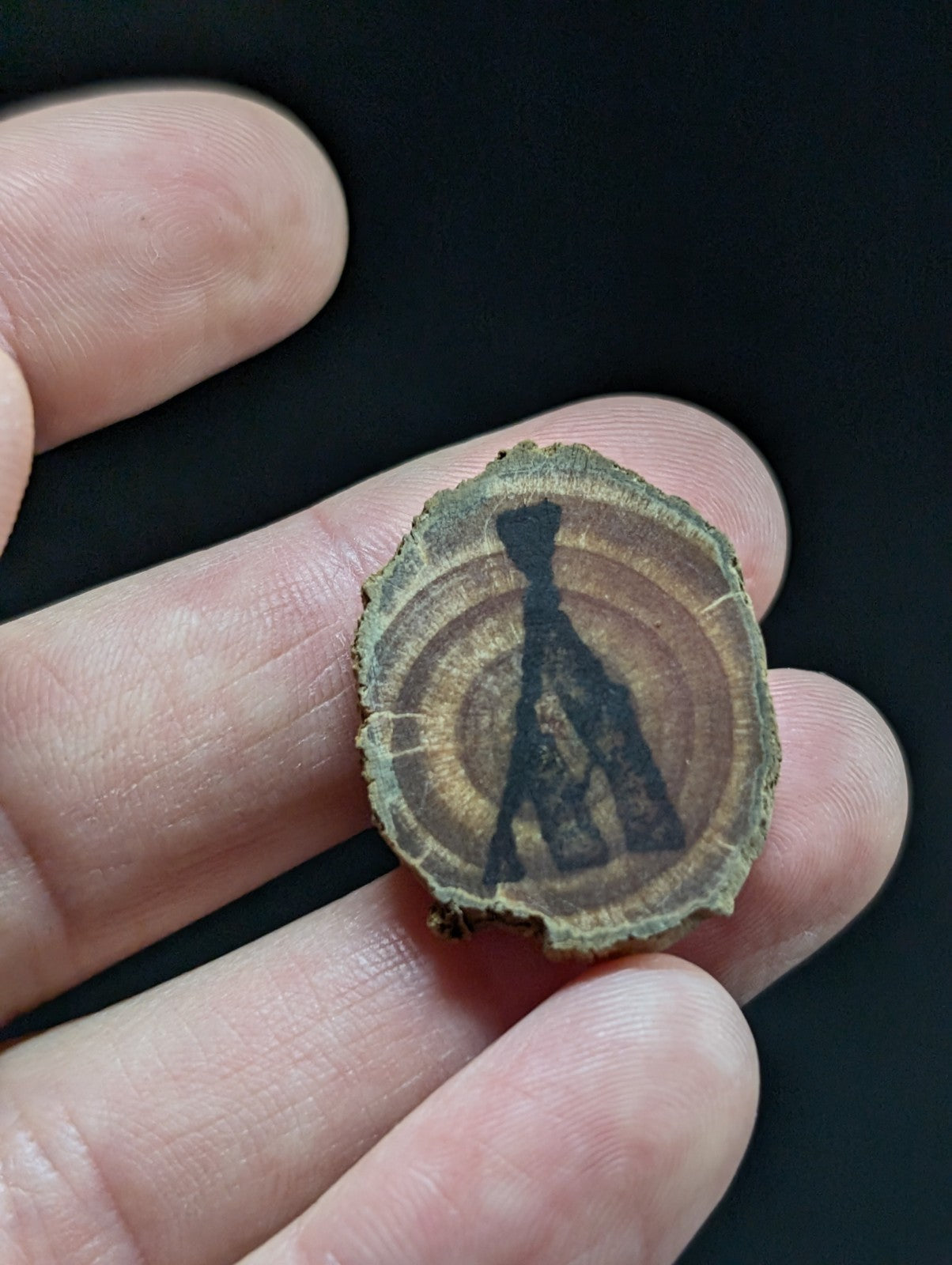 A circular wooden slice with a dark, stylized silhouette of a figure etched into its natural grain.