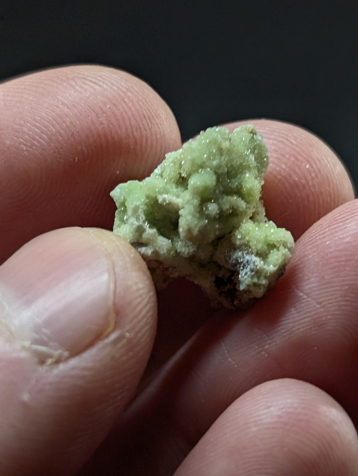 A small, rough-textured green mineral specimen with a crystalline surface and some darker inclusions.