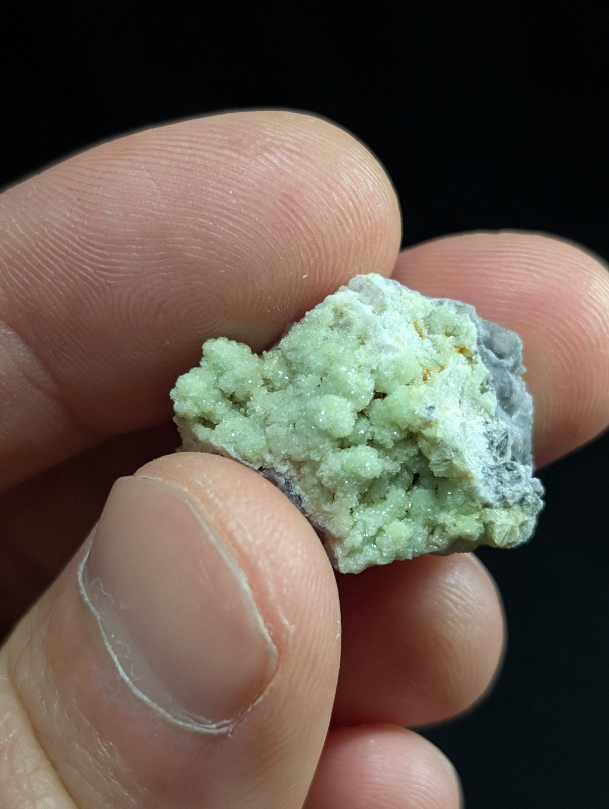 A pale green, crystalline mineral specimen with a rough, textured surface and some grayish matrix.