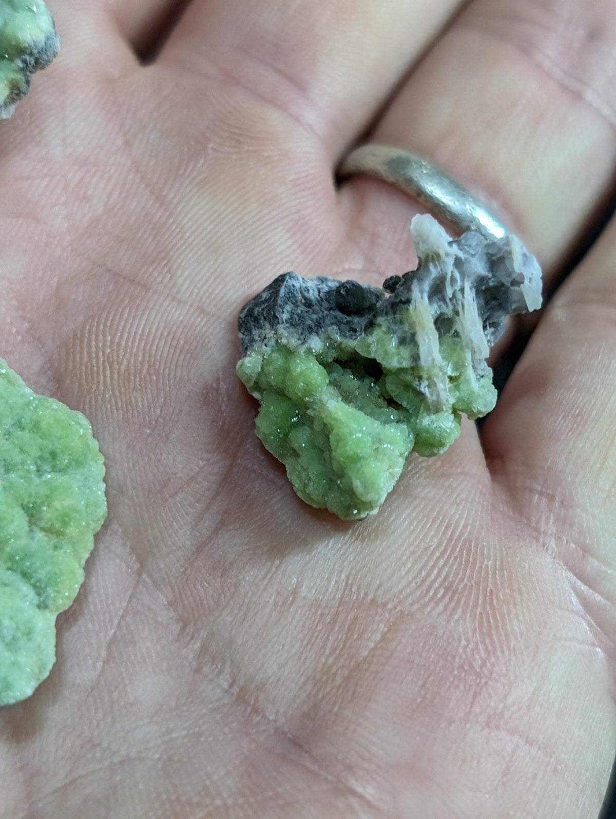 A rough, irregularly shaped green mineral specimen with patches of dark gray and white crystalline formations.