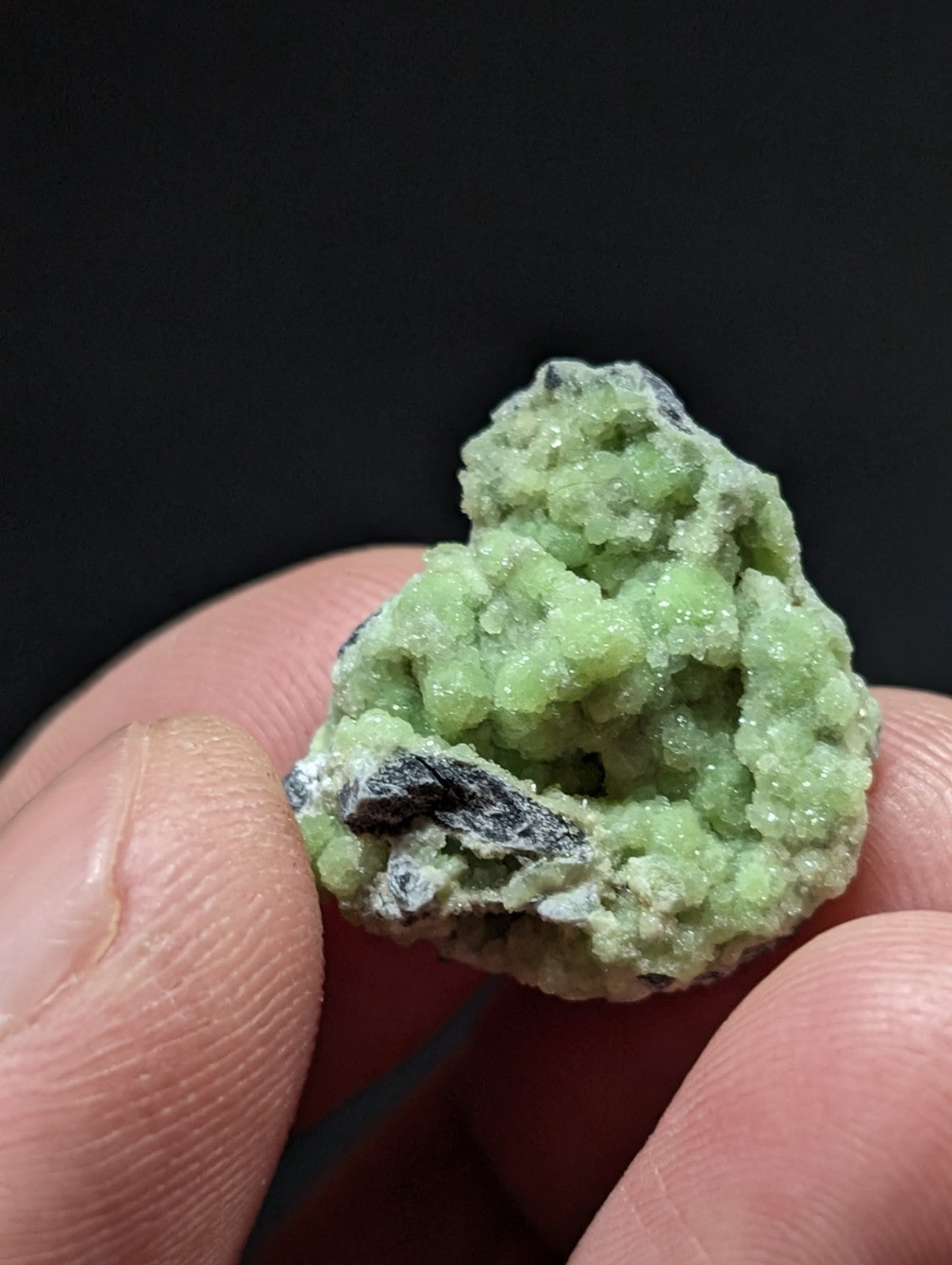 A small, irregularly shaped green mineral specimen with a crystalline texture and dark grayish-black patches.