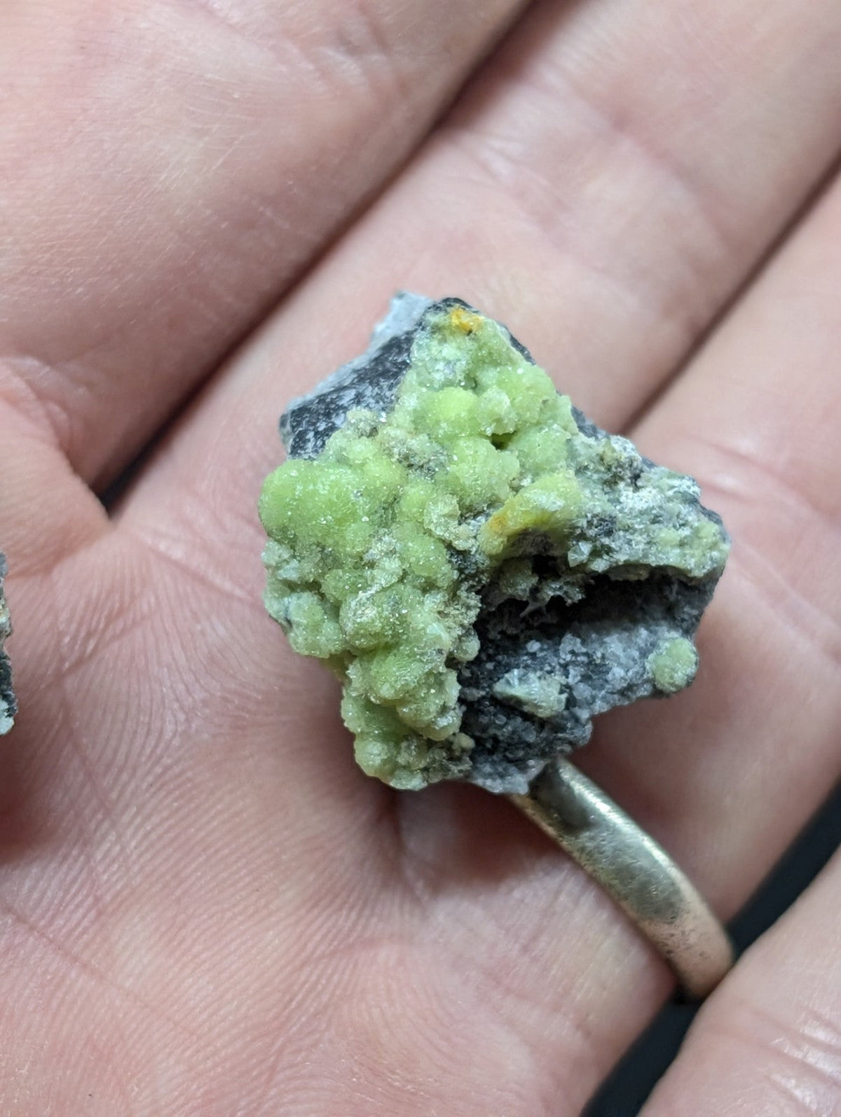 A hand holds a rough, irregularly shaped mineral specimen featuring vibrant lime-green crystalline formations growing from a dark gray matrix.