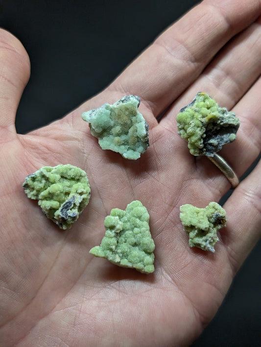 A hand holds five irregularly shaped, light green mineral specimens with dark gray and black patches.