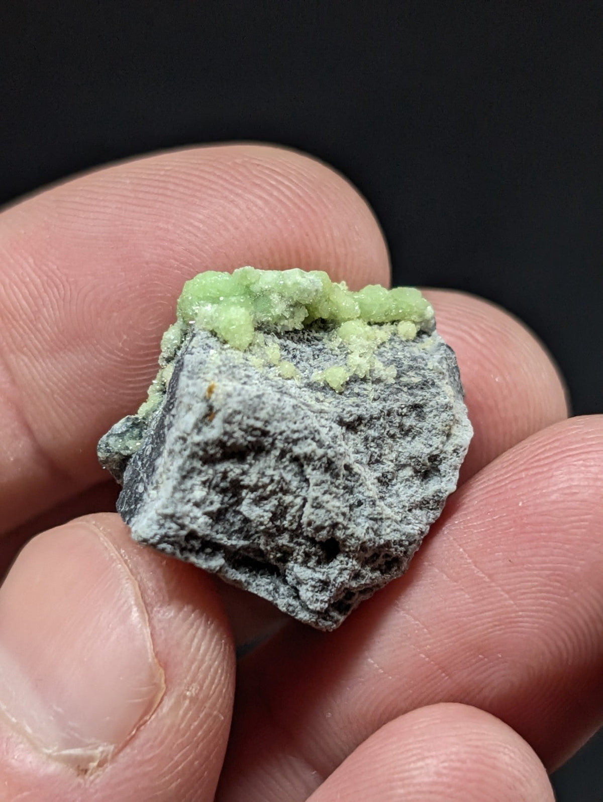 A hand holds a small, rough-textured mineral specimen with a dark gray base and vibrant green crystalline growths on top.