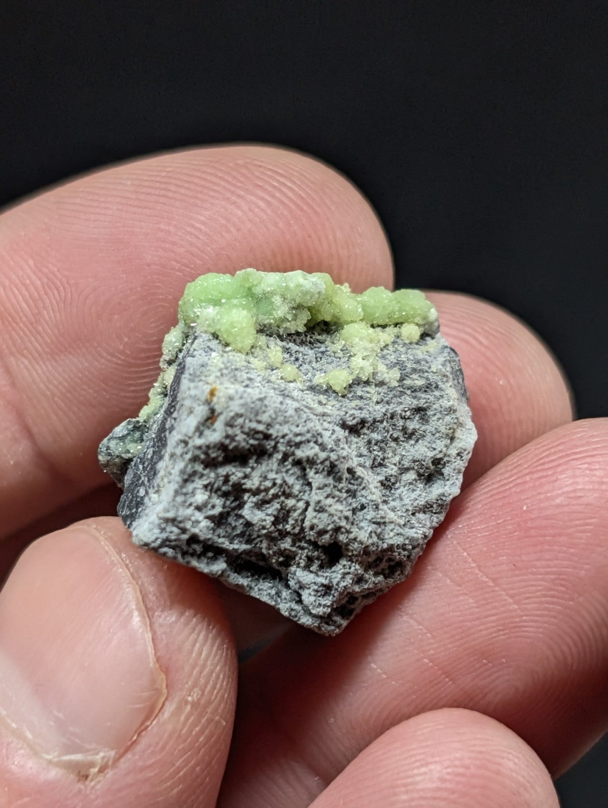 A hand holds a small, rough-textured mineral specimen with a dark gray base and vibrant green crystalline growths on top.